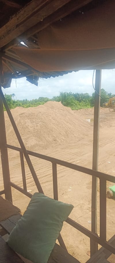 The image presents a view of an outdoor sand processing or storage area within Pontianak, Indonesia, observed from a shaded, elevated structure. The foreground features an overhead wooden beam structure with a torn, light brown fabric or tarp covering, from which several thin wires or ropes descend. A dark brown metal railing with horizontal bars and vertical posts traverses the lower-mid foreground. Resting on a dark wooden surface in the lower-left corner is a single dark green rectangular pillow.
Beyond the railing, the midground is dominated by multiple large, irregularly shaped mounds of light brown sand. The sandy ground exhibits numerous parallel tire tracks. A small, shallow puddle of water is visible on a flat sandy area to the right of the central sand pile. In the far right midground, a segment of a yellow heavy construction vehicle, likely an excavator, is partially visible. The background consists of dense green tropical foliage, including prominent palm trees, under a sky with significant cloud cover. No individuals are present in the scene, and no overt actions or interactions are depicted.