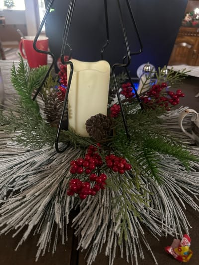 A black metal candle holder is adorned with holiday greenery. The arrangement includes snow-flocked pine needles, bright green fir sprigs, clusters of vibrant red berries, and pine cones. A warm white, battery-operated candle sits within the holder. A red coffee mug peeks out from below. In the upper right, a piece of brightly colored candy wrapping lies on a brown surface.