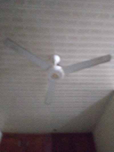 A blurred image captures a three-bladed ceiling fan against a textured ceiling. The fan appears white or light-colored, with the central motor housing also being a similar hue. Below the fan and ceiling, the lower portion of the image is considerably darker, possibly showing a section of a wall and some furniture, but details are indiscernible due to the blur. The photograph has poor focus and exhibits a grainy quality.