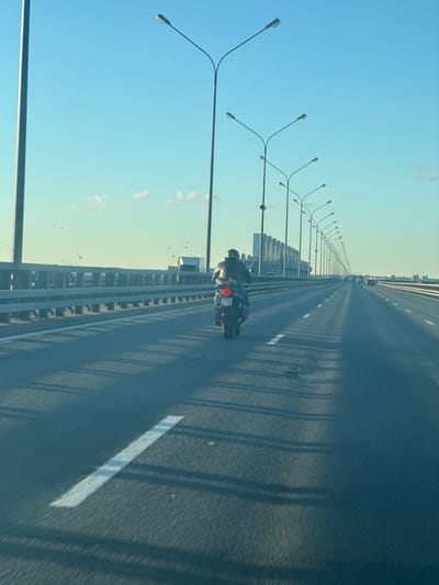 The image shows a view from a vehicle traveling on a highway bridge, likely in or near Kronstadt, Russia, based on the context. The highway has multiple lanes and is lined with streetlights. A motorcycle with a rider wearing a helmet is in the foreground, moving in the same direction. The rider is wearing a dark jacket and blue jeans. The motorcycle has a license plate with the numbers "2288 AT 78". In the distance, other vehicles can be seen, as well as some buildings and industrial structures. Birds are flying in the clear blue sky. The sun is low in the sky, casting long shadows across the road. The environment appears to be an elevated roadway with barriers on either side.
