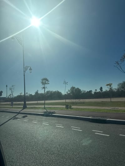 This image depicts an outdoor scene in Ouahat Sidi Brahim, Morocco, under a clear, bright blue sky with a prominent sunburst in the upper left quadrant. The scene is viewed from a low perspective, likely from inside a vehicle.
In the foreground, a dark asphalt road occupies the lower portion of the frame, marked with three sets of parallel white dashed lines on the right side, indicating a lane or shoulder. To the right of the road, a concrete curb separates it from a paved pathway, which is followed by a strip of green grass. Beyond the grass, a lighter-colored dirt or gravel path is visible, with sparse vegetation.
A tall, dark streetlamp, featuring two ornate, cage-like lanterns with decorative lattice work, stands prominently on the left side of the image, extending from near the bottom to the upper left. The streetlamp casts a long, dark shadow diagonally across the road and the adjacent green space to the right.
Several young, sparsely leafed trees are planted intermittently along the grassy median, also casting distinct shadows. In the mid-ground and background, a dense line of diverse trees, including several tall palm trees, forms a green barrier or forested area. No individuals, animals, or moving vehicles are present in the visible portion of the scene. A single metal grate is observed in the curb in the lower right.