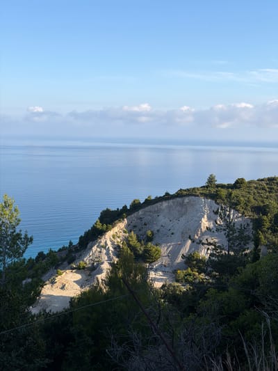 The image depicts a coastal landscape in the Municipal Unit of Sfakiotes, Greece. A steep, light-colored cliff face is visible on the right, with green vegetation growing on its top and along its slopes. The cliff descends toward a body of water, likely the sea, which fills the majority of the left side of the image. The sky is light blue with some scattered clouds. The perspective is from an elevated position.