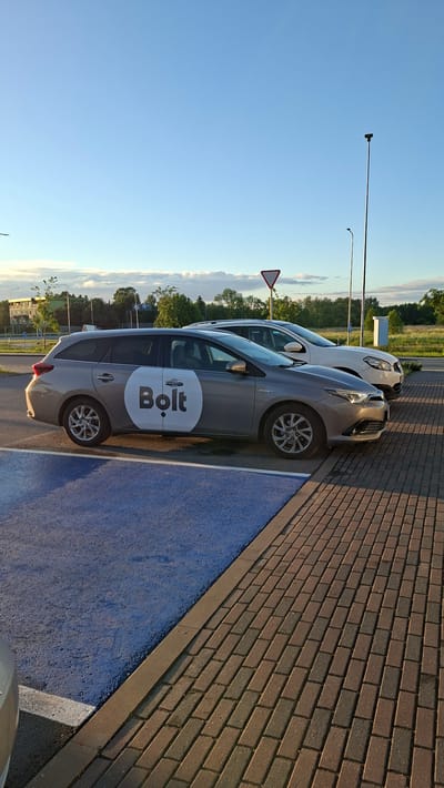 A grey hybrid hatchback with a white circular logo containing the word "Bolt" in black text is parked in a paved parking lot. The car is positioned to the left of a white SUV. A blue painted area is visible in the foreground to the left, and a brick-paved pathway runs along the right side of the parking lot. In the background, trees and buildings are visible under a clear blue sky with some scattered clouds. The scene appears to be a sunny day, possibly late afternoon or early evening, with shadows cast on the ground. The location is identified as Tiskre, Estonia.