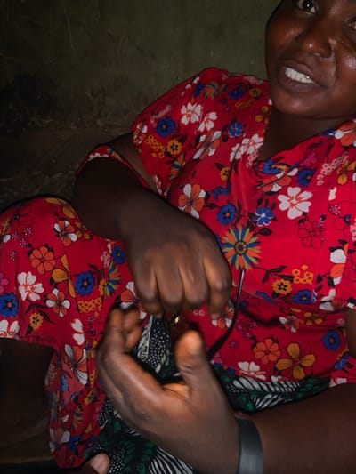 In Project, Nigeria, an individual wearing a red floral-patterned dress and a black wristband is seated against a dark, textured wall. Their hands are clasped in front of them, holding a dark object that appears to be a pair of glasses. The individual's face is partially visible, showing dark skin, a wide smile, and visible teeth. The lighting is dim, with a spotlight effect on the person's face and hands, suggesting an indoor setting or nighttime environment.