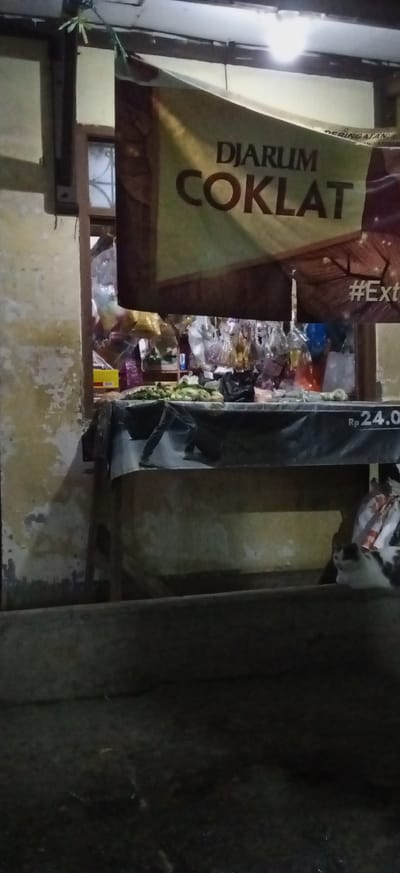 The image captures a dimly lit street stall or small shop, likely operating during the evening or night in Banjaran, Indonesia.
A prominent banner hangs above the counter, featuring the text "DJARUM COKLAT" in large, dark lettering on a lighter background. Partially visible behind it is another banner with the word "PERINGATAN" (warning). To the right of the main banner, a fragment of a hashtag, "#Ext," can be seen.
Below the banners, a counter or table is covered with a dark, possibly plastic, cloth. On this surface, various fresh green produce items, such as vegetables or small fruits, are laid out for sale. Behind the counter, shelves hold numerous small items, many packaged in clear plastic bags, creating a bustling display of goods. A yellow box is also visible further back on a shelf.
To the right side of the counter, a price tag displays "Rp 24.0," indicating a price of 24,000 Indonesian Rupiah. A person's legs and feet, clad in dark trousers and shoes, are partially visible on the dark counter covering, suggesting an individual is standing or working behind the stall.
In the lower right corner of the image, a white and black domestic cat is resting or sitting on a lower ledge, looking towards the left. The background features an aged, textured wall, possibly light yellow or beige, showing signs of wear and a wooden window frame. The overall scene is illuminated by an artificial light source, creating distinct shadows and highlights.