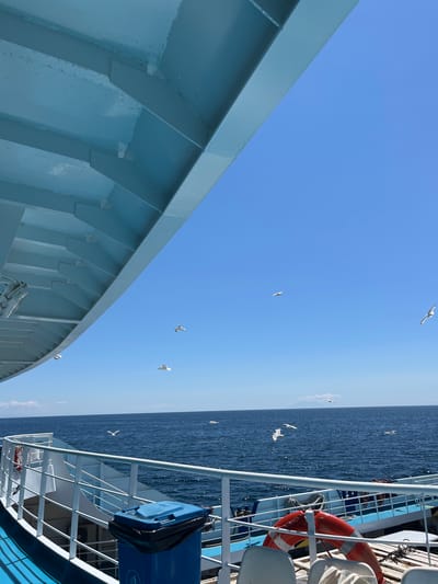 On a bright, sunny day, the perspective is from the deck of a ferry boat looking out towards the Aegean Sea. The ferry's light blue undercarriage with visible structural beams dominates the upper left quadrant of the frame. Below this, the railing of the ferry deck, painted white with a blue trim, runs horizontally across the lower portion of the image. A blue trash bin with a black liner is visible on the deck. Several white seagulls are captured in flight against a clear, vibrant blue sky. The sea is a deep blue, with small whitecaps indicating a gentle breeze. The horizon line is sharp, with a faint outline of land or islands visible in the distance. The overall scene conveys a sense of open space and travel. It is not possible to determine the specific location within the city of Thassos, Greece from this image alone.
