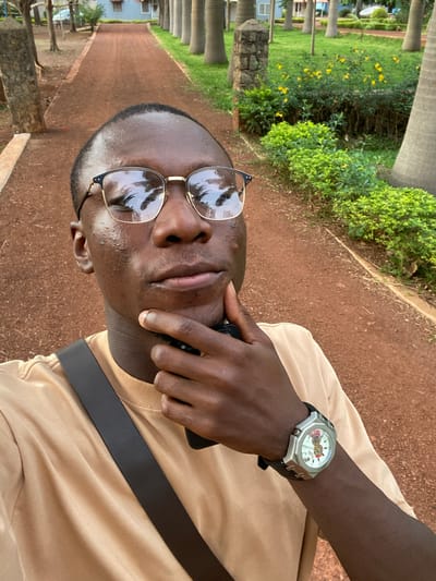 The photograph depicts a man outdoors in Miango, Nigeria. He is wearing glasses, a t-shirt, a watch, and a black strap across his chest. He is holding a phone to his face. The background includes greenery, trees, and a red-brown pathway.