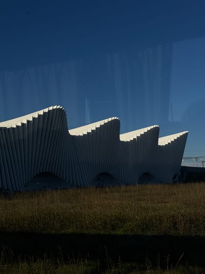 The image displays the Reggio Emilia AV Mediopadana Station in Reggio nell'Emilia, Italy, under a clear blue sky. The station's prominent architectural design features a series of four distinct, light-colored, wave-like segments, each constructed from numerous vertical, angled panels creating a pleated effect. Strong shadows are cast into the inward-facing recesses of these panels, contrasting with the illuminated outer edges. Several arched openings are visible along the base of the structure. The foreground is composed of a field of dry, light brown and green tall grass, with a dark horizontal shadow band spanning the immediate bottom edge of the frame. A small, dark, indistinct object is present in the upper left quadrant of the clear blue sky. Faint vertical reflections are visible across the upper portion of the image, indicating the photograph was captured through a window. Partially visible indistinct industrial or utility structures are present on the far right horizon, behind the station building. No human figures are present.
