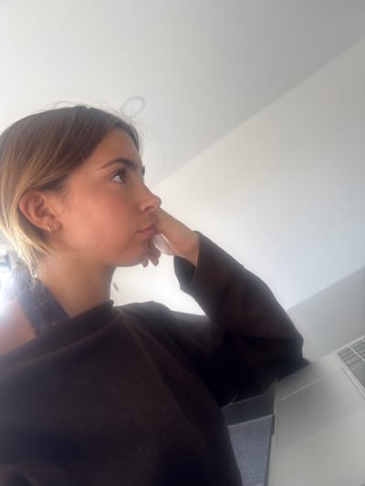 The image shows a woman sitting indoors, angled upwards. She has light brown hair with blonde highlights, fair skin, and is wearing a brown sweater. Her right hand is touching her face near her mouth. She is looking upwards, towards a light source. A laptop is partially visible to the right of the image. The background is a plain white wall. There is no information given to suggest that the image was taken in Torres Vedras, Portugal.