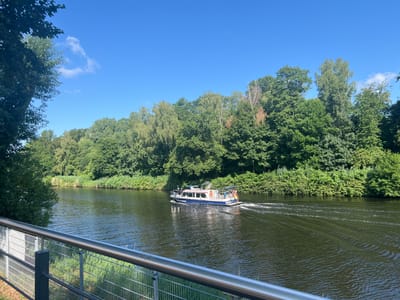 A police boat, identified as "SCHIFFAHRTSPOLIZEI BERLIN," travels east on the Teltow Canal in Teltow, Germany. The boat has a blue hull with white and red accents and a German flag displayed at the stern. The canal is surrounded by lush green trees and vegetation on both banks. A metal railing and fence line the foreground of the image, suggesting a public walkway or embankment. The sky is clear blue with a few wispy clouds. The water is dark and calm, with ripples trailing behind the moving boat.