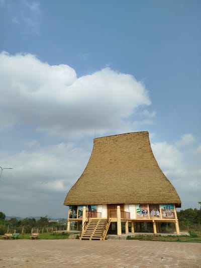 The image depicts a traditional house in Đà Giang, Vietnam. The house has a tall, thatched roof and is elevated on wooden posts. The walls are painted with murals depicting people in traditional clothing. There are wooden steps leading up to the entrance. The house is located on a brick paved area with a few benches nearby. The background includes trees and a partly cloudy sky.