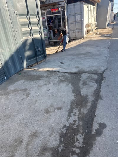 A man, wearing a black t-shirt and blue jeans, is sweeping the ground in front of a row of shipping containers in Kara-Suu, Kyrgyzstan. He is holding a broom and appears to be in the process of cleaning the concrete area. The shipping containers are grey and appear to be used as small shops or stalls. The ground is a mix of concrete and asphalt, with some visible water stains and cracks. There are other shipping containers in the background, and a street stretches out behind them. The overall scene depicts a commercial area with a focus on a single individual performing a cleaning task.
