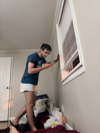 The original image, rotated 90 degrees clockwise, presents an indoor scene where a man appears to be horizontally suspended or lying down, interacting with a ceiling fixture. A young man with dark hair, a short beard, and glasses is the central figure, wearing a dark blue t-shirt and white shorts. He is reaching with his right hand into a rectangular air vent or return located in the light-colored ceiling. He holds a white smartphone in his left hand, seemingly using it to illuminate the area or for reference.
On the far right of the frame, partially visible, is a hand and forearm clad in a maroon long-sleeved garment, belonging to another person who seems to be standing nearby. In the background, what would be a white door with a silver doorknob and a key inserted is seen. Adjacent to this door, a casual pile of clothes or fabric in various colors (white, blue, maroon) is noticeable. The walls are a light, neutral color, suggesting a typical residential room.
The man appears focused on the task involving the ceiling vent, possibly inspecting or repairing it. There are no strong emotions visible. The time of day and weather conditions cannot be determined from the indoor setting. No visible text is present in the image. The scene is set in a residential interior within Franklin Township, United States of America.