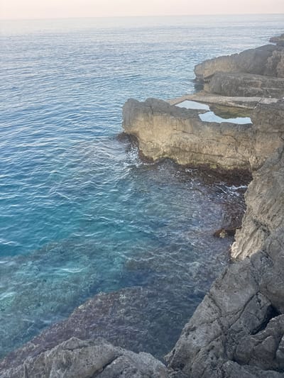The image depicts a rocky coastline with clear turquoise water lapping at its base. The rocks are weathered and appear to have formed natural pools or platforms at different elevations. In the distance, the calm sea meets a hazy horizon under a pale sky. The scene is devoid of any people, structures, or significant activity, presenting a serene natural landscape. The location is identified as Višnjevo, Montenegro.