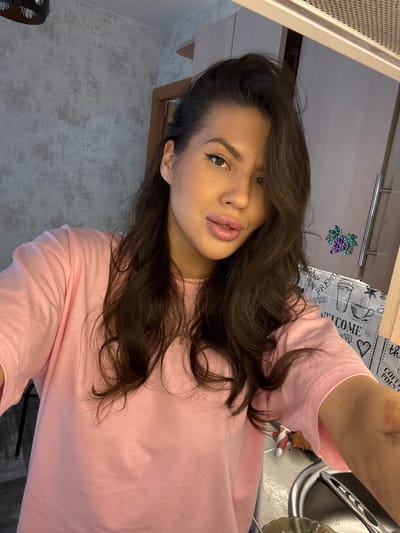 An indoor selfie captures a female subject, likely in a kitchen or domestic setting within Solnechnyy, Russia. The subject, a woman, has long, dark brown, wavy hair and is wearing a plain pink short-sleeved t-shirt. Her facial features include defined eyeliner on both eyes and full lips. A noticeable bruise, exhibiting purple and yellowish-green discoloration, is present on her right forearm.

The background to the upper left features light gray wallpaper with a textured, mottled pattern, and a portion of a dark-colored light fixture is visible. To the right and behind the subject, a wooden door frame is partially visible. Further right, a kitchen area is depicted, including a stainless steel sink with a faucet and a partial view of metallic utensils within a dish rack. On the counter, a red and white kitchen tool handle and a piece of food item, possibly bread or pastry, are present. Above the counter, a backsplash with a repeating grey and white coffee-themed pattern, featuring illustrations of coffee cups, text like "WELCOME" and "COFFEE POINT," is visible. A magnetic decoration shaped like a cluster of purple grapes with green leaves is affixed to a metallic surface above the backsplash. Adjacent to this decoration, a vertical metallic handle of a cabinet or appliance is visible. A metal grate structure, consistent with a ventilation hood or overhead vent, spans the upper right portion of the image. The subject is directly facing the camera, indicating a self-portrait.