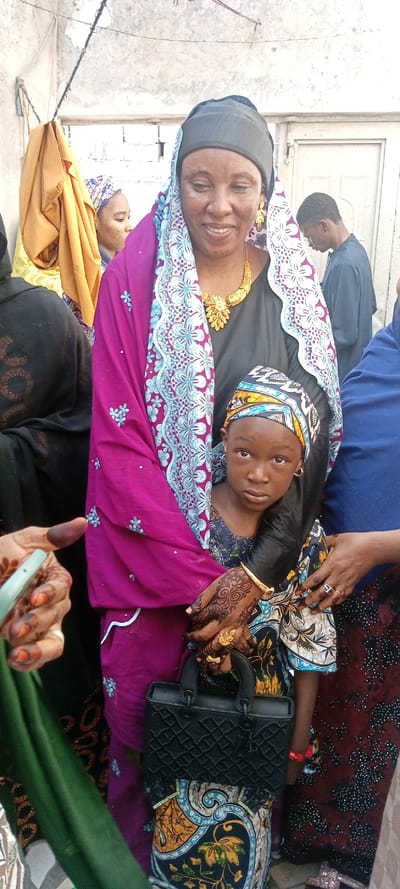 The image depicts a group of individuals in an outdoor setting within the city of Dan Maliki, Nigeria.
In the foreground, an adult female with dark skin occupies the central frame, smiling and looking slightly to her left. She wears a black head covering underneath a vibrant magenta headscarf adorned with light blue floral embroidery and a wide patterned border. A gold-colored ornate choker-style necklace with a prominent central floral motif and matching leaf-shaped dangle earrings are visible. Her left arm supports a young child, who stands in front of her. This child, also with dark skin, wears a multi-colored patterned headwrap (yellow, blue, black, white) and small silver hoop earrings, looking directly at the viewer with a neutral expression. The child's right arm displays a gold-colored bangle bracelet. Both the adult woman's left hand and the child's right hand grasp a black, textured handbag with a top handle. The adult woman's left hand is intricately decorated with dark henna patterns and displays gold-colored rings.
To the immediate left, a partially visible female figure wears a dark garment with circular patterns. Her right hand, also adorned with henna and with darkened fingertips, holds a light green, elongated object. Further left and slightly behind the central adult, another female figure with a patterned headwrap is partially obscured.
In the mid-ground to the right, a male individual with dark skin, dressed in a dark blue or black long-sleeved garment, is visible in profile, looking downwards and away from the camera. A female hand with a ring featuring a light-colored stone is partially visible on the far right.
The background consists of a light-colored, textured concrete or plastered wall, showing minor discoloration and surface variations. A horizontal bar or wire is present in the upper left corner, from which a piece of light brown fabric hangs. A framed opening, possibly a window, is visible in the upper left, and a white door or doorway is present in the upper right. The scene is illuminated by bright, natural daylight.