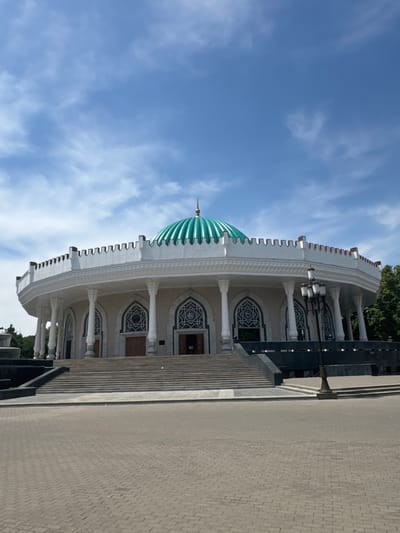 The image depicts the Id Kah Mosque in Kashgar, Uzbekistan, viewed from a low angle. The mosque features a large, green, ribbed dome and an arcade with white columns and arched windows with intricate geometric patterns. The building has multiple stories, and a wide set of concrete steps lead up to the entrance. The exterior is white with some darker trim. To the right, there is a paved area with a brick pattern. The sky is blue with scattered clouds.