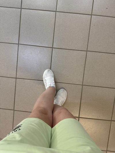 A first-person perspective shot shows a person's legs from the knees down, wearing light green shorts and white slip-on sneakers. The individual is standing on a floor tiled with square, light beige tiles. The scene is indoors, likely on a tiled surface, possibly a public building or walkway. The image does not provide any specific visual cues to pinpoint a location within the city of Inzer, Russia.