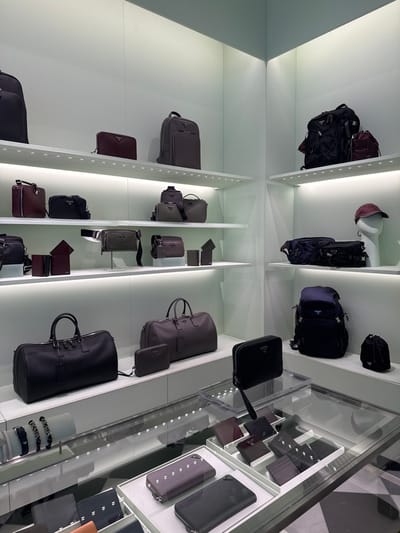 The image depicts the interior of a Prada retail store, presumably located within Charles de Gaulle Airport in Roissy-en-France, France. The space features brightly lit white walls and shelving units arranged in a corner display. Numerous Prada leather goods are meticulously arranged on these shelves and on glass-covered counters.
Displayed items include a variety of bags: several backpacks in black, dark grey, and dark blue; large duffle bags in black and dark grey; smaller shoulder, crossbody, and belt bags in shades of black, maroon, dark olive, and grey; and a small black drawstring bag. A white mannequin head wearing a maroon baseball cap is visible on an upper shelf to the right. The lower section of the store features glass display cases showcasing wallets and small leather goods in black, maroon, and dark purple hues, some elevated on clear acrylic risers. Silver-toned bracelets are displayed on a white stand within a glass case on the left. The visible flooring, seen through reflection, features a black and white geometric pattern. No individuals are present in the scene.
