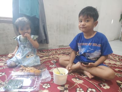 Two children are sitting on a red carpet with a floral pattern, in what appears to be a domestic indoor setting in Cintadamai, Indonesia. A younger child, a girl, is on the left, wearing a light blue dress with a cat print, and she is bringing food to her mouth. To her right, an older child, a boy, is sitting cross-legged, holding a smartphone in his right hand. In front of the children is a clear plastic container with various fried snacks, and a white paper cup with a spoon. A plastic bag is also visible near the cup. The background shows a wall and what looks like clothing hanging on a rack. The lighting suggests it is daytime.