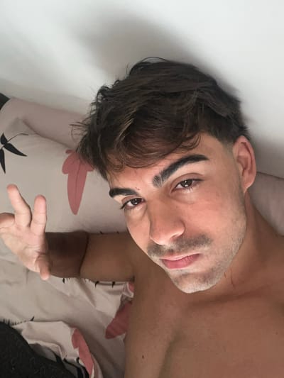 A portrait-style photograph depicts a young man, likely in his 20s or 30s, lying down in what appears to be a bed. He has dark, slightly dishevelled hair, prominent dark eyebrows, and dark eyes that gaze directly at the viewer with a thoughtful or curious expression. His face has light stubble. The man's upper torso is bare, revealing his skin, and a faint scar is visible on his right shoulder/neck area.

He is positioned with his head slightly tilted, resting on or near a white pillow adorned with a decorative pattern of pink and black stylized leaves. The bedding below him exhibits a similar leaf motif. A plain white wall serves as the background behind his head and to his right. His right hand is partially visible in the foreground, extended towards the camera with fingers gently curled. The lighting is soft and diffused, creating an intimate and casual atmosphere, possibly indoors in Sarandí, Argentina. No text is visible in the image.