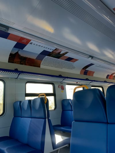 The image shows the interior of a train carriage. Blue upholstered seats with white and grey trim are visible in the foreground and middle ground, arranged in rows. Behind the seats, a window looks out onto a sunlit exterior, though the view is obscured by reflections. Above the window, a blue and orange banner is affixed to the ceiling, featuring text in Cyrillic script and images of buildings and landscapes. The ceiling itself is white and perforated. A yellow handle is attached to the back of a seat near the window. The lighting suggests it is daytime. The text on the banner includes the word "КАМЕНСКАЯ" and mentions "Ландшафтный парк «Лога»" which translates to "Landscape Park 'Loga'". According to information on the banner, this park is located in the city of Kameshkovo, in the Vladimir region, Russia. However, the prompt specifically asks for the location of Kolesnikovo, Russia. Based on the visible information, this is a scene within a train and the banner describes the "Loga" Landscape Park. The prompt's mention of Kolesnikovo is not supported by direct visual evidence in the image.