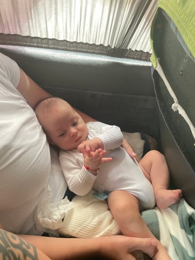 A newborn baby is lying on a blanket, held by an adult whose arm is visible on the left side of the image. The baby is wearing a white bodysuit and has a pacifier. The adult has a tattoo on their arm. The scene appears to be indoors, possibly in a vehicle given the presence of a dark-colored seat and window coverings in the background. The image quality suggests it was taken with a mobile device. There is no specific information in the image to indicate the location is in Răuțel, Moldova.