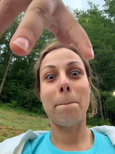 A close-up, slightly distorted selfie is captured from a low angle, featuring a young woman's face. Her expression is playful, with pursed lips and wide, curious eyes. A hand, partially obscured by the foreground, is reaching towards the camera, its index finger and thumb prominently displayed.

The woman is wearing a bright blue t-shirt, and a white hooded garment is draped over her shoulders. Her hair, a light brown hue, is slightly messy and appears to be caught by a breeze. The background is filled with lush green trees, suggesting an outdoor setting. The lighting indicates it is daytime, with soft, diffused light illuminating the scene.

The image appears to be taken in Novo Bardo, Bulgaria. The natural, wooded environment combined with the casual attire of the subject points towards a relaxed, possibly recreational outing. No visible text is present in the image. The overall mood is lighthearted and spontaneous.