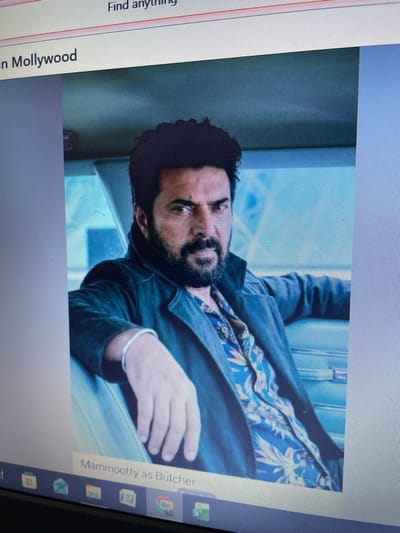 The image captures a digital photograph displayed on a computer screen, located in Noida, India. The primary subject on the screen is a close-up portrait of an adult male, positioned centrally and looking directly forward with a serious, intense expression. He possesses dark, wavy hair and a full beard. The man is attired in a dark, possibly denim or canvas, jacket worn over a blue shirt featuring a light-colored, possibly beige or white, leaf or floral pattern. A single, plain silver-colored bangle is visible on his left wrist. He appears to be seated within the interior of a vehicle, suggested by the light blue-grey paneling visible in the background. Above the featured photograph, text on the screen reads "in Mollywood" and a "Find anything" search bar is present. Below the photograph, the text "Mammootty as Butcher" is displayed. The lower edge of the computer screen reveals a partial view of a Windows operating system taskbar, showing various application icons.