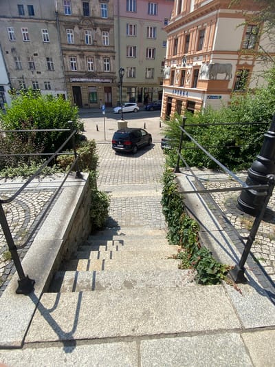 In Świdnica, Poland, a black Volkswagen Touran is parked on a cobblestone street at the bottom of a wide, stone staircase. The staircase is bordered by stone walls and metal railings, with green ivy growing along the lower sections of the wall. The street is lined with old European buildings, one of which has a sign reading "APTEKA" (pharmacy) and decorative relief sculptures of bulls above the entrance. A white Volkswagen Golf is parked further down the street. The scene is illuminated by bright sunlight, casting distinct shadows on the staircase and street.