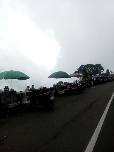 A row of motorbikes parks along a dark asphalt road beside a low concrete barrier. Several green umbrellas shade small makeshift food stalls set up behind the barrier. Patrons are present at some of the stalls, suggesting the area is a rest stop or viewpoint. The sky is overcast with heavy, diffused light, and a tree provides a backdrop to the scene. A white painted lane divider runs alongside the road.