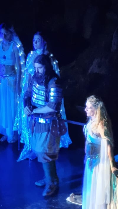 The image depicts a dimly lit indoor scene, possibly a stage production, with several individuals in elaborate costumes. In the foreground, a man with a long beard and dark hair, wearing a segmented chest piece and leather armor, holds what appears to be a sword. To his left, a woman in a white, flowing garment decorated with strings of lights stands, her face turned away from the camera. Further left, another woman, also in a white costume with similar light embellishments, is partially visible. To the right of the man in armor, a woman with blonde hair, wearing a silver headdress and a light blue and white draped costume, is also facing away from the camera, her gaze directed downwards. The floor appears to be a dark, reflective surface, likely a stage. The overall ambiance is theatrical, with a focus on costumes and dramatic lighting, which casts a blue hue across the scene. There is no specific indication of the location being in Sant Joan les Fonts, Spain, within the provided image.