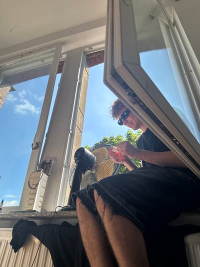 A young adult with curly hair and sunglasses sits on a windowsill in Hamm, Germany, in front of an open window. They are wearing a black t-shirt and ripped black denim shorts. The person is holding a small, pink object in their hands. A black electric fan is positioned behind them. On the windowsill, there are two bottles of Moët & Chandon champagne and a framed picture. Outside the window, a blue sky with scattered clouds and green trees are visible. The scene appears to be indoors, near a radiator covered with a black garment.