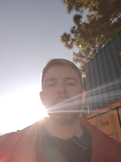 The image presents a low-angle, close-up selfie of an adult male, taken outdoors. The subject's face, partially obscured by severe lens flare originating from the lower-left, shows short brown hair, visible stubble, and direct gaze into the camera. He wears a dark-colored shirt and a red outer garment draped over his shoulders. A black rectangular device, possibly a microphone or small electronic accessory, is clipped to the collar of his dark shirt. The background, largely visible in the upper right, features coniferous tree foliage and two stacked shipping containers. The upper container is dark blue or grey, while the lower one is orange or yellow, bearing the stenciled alphanumeric designation "UXJH". The sky is clear and light blue. Strong direct sunlight from the lower left creates prominent lens flares. The specified location is the city of Nesher, Israel.