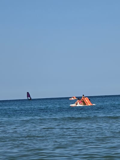 The image depicts a clear, sunny day at a coastal location, likely Ahtopol, Bulgaria, with a vast expanse of blue sky meeting a tranquil blue sea. In the distance, a lone windsurfer navigates the water, their purple sail a striking contrast against the horizon. Closer to the foreground, two paddle boats are visible. One paddle boat carries a group of people enjoying a leisurely outing. Another paddle boat is occupied by a family; a man and a child are seated on the edge, while a woman and a young girl are on the slide, with the girl in a pink swimsuit. The water is calm with gentle ripples, suggesting a peaceful day at the beach.