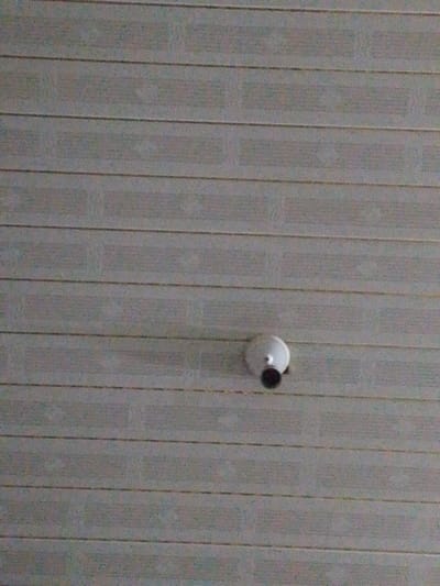 The image shows a white security camera mounted on a ceiling with horizontal planks. The camera has a round base and a dark, possibly infrared, lens. The ceiling appears to be made of light gray or white planks, with visible seams or lines between them. The planks run horizontally across the frame, and the camera is positioned slightly below the center. The lighting is somewhat dim and flat, creating soft shadows.