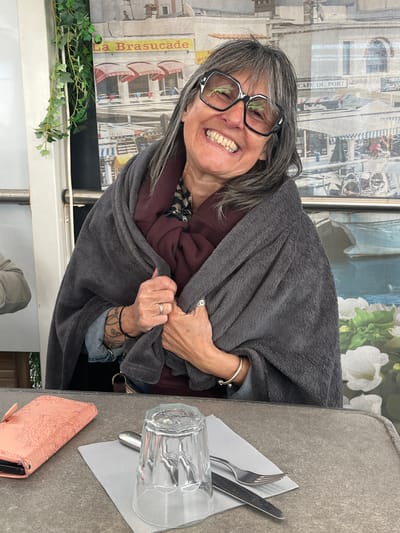 An older woman with grey hair and dark-rimmed glasses smiles broadly at the camera, her teeth visible in a joyful expression. She is wrapped in a cozy, grey fleece blanket or shawl, which covers her dark red top. A silver bracelet adorns her left wrist, and a silver ring is visible on her finger. A black tattoo with swirling patterns can be seen on her inner right forearm, peeking from under a denim-like sleeve.

She appears to be seated at a grey, textured table at what looks like an outdoor or semi-outdoor cafe or restaurant in Palavas-les-Flots, France. On the table to her right are an empty clear glass, a folded light grey napkin, a fork, and a knife. A pink or peach-colored phone case with an intricate embossed floral or mandala pattern rests nearby.

The background, visible through a window or glass partition behind her, features a decorative screen or curtain. This screen depicts stylized buildings, possibly a harbor scene. Partially visible text on the screen reads "CAFE DE PORT" and, vertically, "La Brasucade," a term associated with Southern French cuisine, further supporting the given location. A green, leafy vine (possibly ivy) is draped along the bottom left edge of the frame, adding a touch of natural decor. The lighting suggests it is daytime, and the woman's cozy blanket implies a cool but comfortable atmosphere.