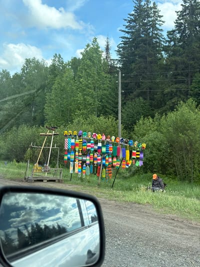 Captured from a vehicle's perspective, the image reveals a roadside scene, likely in a rural or semi-rural setting, though Черёмушки, Russia, is not discernible. The scene features a display of handcrafted items, possibly bath loofahs or similar objects, arrayed on a metal rack along the roadside. They are colorful and varied, some adorned with flowers or patterned designs. Behind the display is a background of lush green foliage, composed of trees and underbrush. An individual wearing an orange hat is visible tending to the grass next to the display. A utility pole with electrical lines runs alongside the road. The road itself appears unpaved and gravelly. The vehicle's side mirror is visible in the foreground, reflecting the sky and surrounding trees.