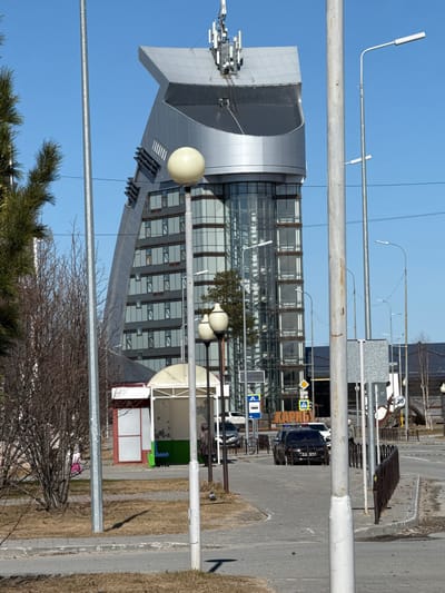 The image shows a modern building in Beloyarsky, Russia, characterized by a combination of silver metal cladding and extensive glass windows. The building has a distinctive, angular design with a notable section that curves. Various antennas and equipment are visible on the building's roof. Street lights are placed in front of the building, obscuring parts of it.
In the foreground, there are trees with bare branches, as well as evergreen trees, indicating a cool climate or early spring. A pedestrian walkway, a bus stop, a parking area, and a road are visible next to the building. A white van and a black vehicle are parked. In the distance, there are indistinct structures and landscape. The sky is clear and blue.