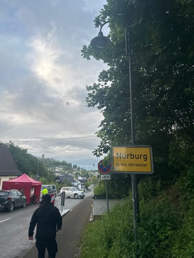The image depicts an overcast outdoor scene in the village of Nürburg, Kreis Ahrweiler, Germany, viewed from a slightly elevated position. In the immediate foreground, a person wearing a dark hooded jacket and dark trousers walks away from the camera on a paved sidewalk. To the left of the sidewalk, an asphalt road runs parallel.

On the left side of the road, a dark grey SUV is parked adjacent to a red pop-up canopy tent. Behind this tent, a dark-colored van or SUV is partially visible, next to which a person in a bright yellow/green high-visibility vest is standing. Further down the road on the right, a silver Mercedes-Benz GLK SUV is visible, and a white sedan is parked behind it.

A prominent yellow rectangular town sign is positioned on the right side of the image, stating "Nürburg" in large black letters and "Kreis Ahrweiler" below. Below this sign, a circular "No Parking" sign (red circle with a diagonal line on a blue background) is visible, with a smaller white rectangular sign beneath it. A black streetlight post with a curved arm and lamp head stands on the right, partially obscured by dense green foliage from trees.

The background features a hilly landscape dotted with numerous residential buildings, characteristic of a village. On the most distant and prominent hill, the stone tower of Nürburg Castle is clearly visible against the cloudy sky. The sky is predominantly grey with patches of brighter light breaking through the clouds. Overall vegetation consists of dense green trees and bushes lining the road and covering the hillsides.