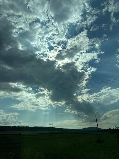 The image captures a dramatic sky over a rural landscape. The sky is filled with a dynamic display of clouds, ranging from dark, heavy formations to wispy, white ones. Rays of sunlight break through the clouds, creating a spotlight effect. Below the sky, a silhouetted horizon features rolling hills covered in trees. In the foreground, a dark green field appears to be a cultivated area, possibly farmland. Several power lines stretch horizontally across the frame, intersecting the sky. A single, tall utility pole stands prominently on the right side of the image, leaning slightly. In the distance, a large electricity pylon is visible. The overall impression is one of natural beauty with a touch of human infrastructure. The image does not depict any people, actions, or interactions. The specific location in the city of Boryana, Bulgaria, is not directly evident from the visual elements alone, but the landscape suggests a rural setting consistent with that region.