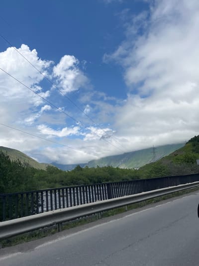 A cloudy sky with bright blue patches is visible above a mountain landscape in the city of Achkhoti, Georgia. Several power lines crisscross the sky. In the midground, a metal guardrail and fence separate a road from a body of water. The water flows through a valley with lush green trees on either side. The background features steep, green mountains, partially obscured by clouds. Power pylons are visible on the slopes of the mountains.