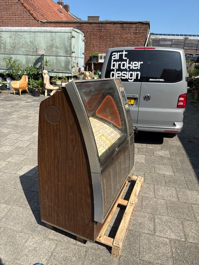 A vintage jukebox sits outdoors on a paved surface, partially on a wooden pallet. Beside it is a gray Volkswagen van with "art broker design" in large white letters on its rear window. In the background, there are buildings of brick and what appears to be a large container or trailer, along with potted plants and a chair. The scene is illuminated by bright daylight.