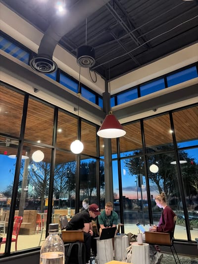 Inside a glass-walled cafe at dusk, people work on laptops. Three are seated, one standing. Outside, bare trees are visible against a fading sky. Above, industrial-style lighting hangs from a high ceiling with exposed ducts. One light fixture features a conical red shade. The cafe appears modern with a blend of natural light and artificial illumination. The image is taken from a tilted perspective.