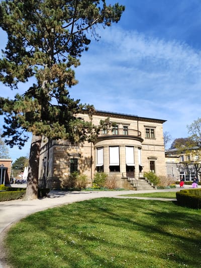 The image depicts the exterior of a three-story sandstone building with a curved facade element. A large pine tree obscures part of the structure. A pathway runs alongside the building, bordering a green lawn. In the background, other buildings and trees are visible. People are present in the far background near the building with the white doors. The sky is blue with streaky clouds. The building is in Bayreuth, Germany.