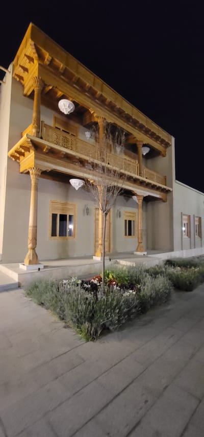 This media file depicts an exterior view of a building at night, likely in Satkak, Uzbekistan, as suggested by the architectural style. The building features intricately carved wooden eaves and decorative elements, suggesting a traditional or culturally significant structure. The scene is illuminated by artificial lighting, casting a warm glow on the wooden architecture and the facade of the building.
The subjects in the image include the building itself, with its visible windows and ornate woodwork. There are also garden beds with foliage and flowers, indicating a landscaped exterior. Cobweb-like structures are visible hanging from the wooden eaves, adding a somewhat eerie or aged aesthetic to the scene. There are no people visible in the image.
The activity taking place is not evident, as the image captures a static scene at night. Notable details include the time of day being night, with a dark sky visible. The weather appears calm and clear, given the absence of rain or wind effects. The architecture suggests a location with a rich cultural heritage, possibly a historic building, a guesthouse, or a cultural center. The overall mood is quiet and somewhat mysterious due to the nighttime setting and the presence of the cobwebs. No visible text is present in the image.