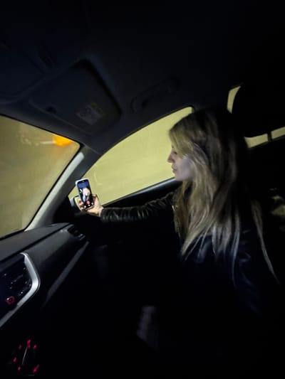 The image depicts the interior of a vehicle, likely positioned within the Sozina Tunnel in Montenegro, indicated by the pervasive uniform yellowish-green light illuminating the exterior visible through the windows. An orange light source is also discernible through the upper left portion of the windshield. A female individual, with long, light-colored hair, is seated in the front passenger seat, wearing a dark jacket. She holds a smartphone in her extended right hand, actively engaged in a video call, with a person's face visible on the illuminated screen. Her profile is visible, and her gaze is directed forward, seemingly towards the phone or the scene outside. The car's dashboard features air vents and control buttons illuminated with red light.
