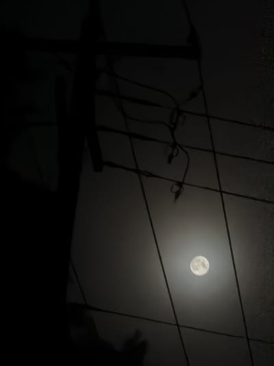 The image is a nighttime photograph of a full moon in a hazy sky. A silhouetted utility pole and power lines are in the foreground, obscuring parts of the moon and sky. The overall scene is dark and moody, with the moon being the primary source of light. The location is the city of Hampton Bays, United States of America.