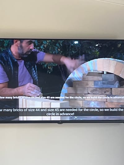 The image shows a close-up of a person's hands and the lower part of their body as they are building a circular structure, likely a pizza oven or fireplace, out of bricks. The person is wearing a purple t-shirt and a black vest. Their hands are positioned as if they are placing or adjusting bricks. The structure is made of stacked bricks of various sizes and colors, with a partially completed arched opening. Behind the person and the structure, there is foliage, suggesting an outdoor setting. The lighting appears to be natural, possibly daylight, with some shadows visible.
Text is visible on the right side of the image, running vertically. It reads: "How many bricks of size 44 and size 45 are needed for the circle, so we build the circle in advance!" This suggests the context is related to construction or DIY, possibly a tutorial or a project documentation. The location context provided is Rosenovo, Bulgaria, but there are no specific visual cues in the image to confirm this. There are no clear indicators of time of day or weather conditions beyond general daylight. The emotion conveyed is one of focused activity and construction.