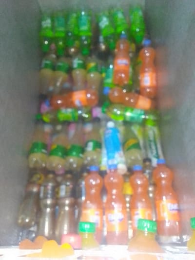 An image of a cooler filled with assorted beverages. Green Sprite bottles occupy the top shelf, followed by rows of yellow-tinted drinks, and a mix of orange Fanta and green soda bottles. The bottom level features an array of drinks, including orange Fanta bottles with blue caps and smaller, green-capped juice bottles. The cooler appears well-stocked, providing a variety of choices.